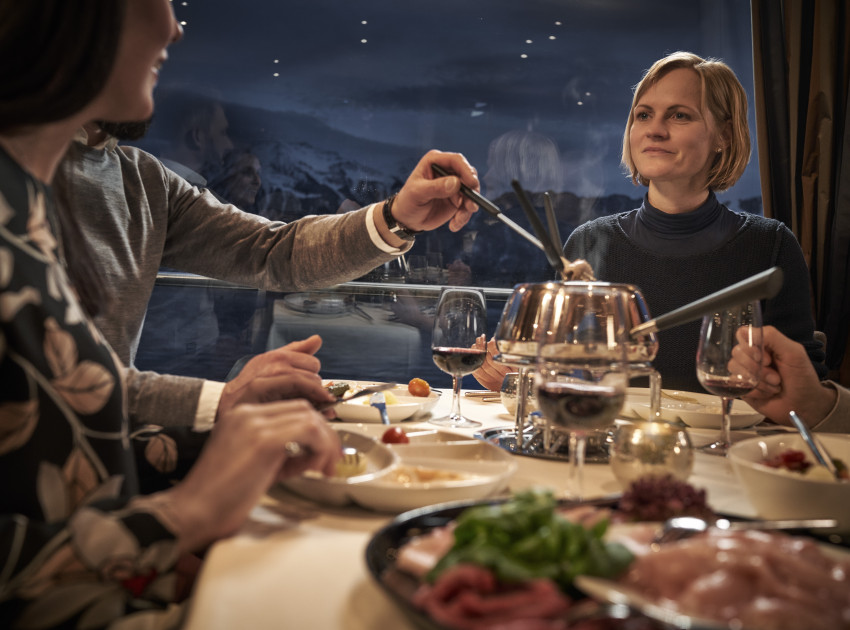 Freunde essen gemeinsam Fondue Chinoise auf dem Schiff und vom Fenster aus hat man Sicht auf die Berge. 